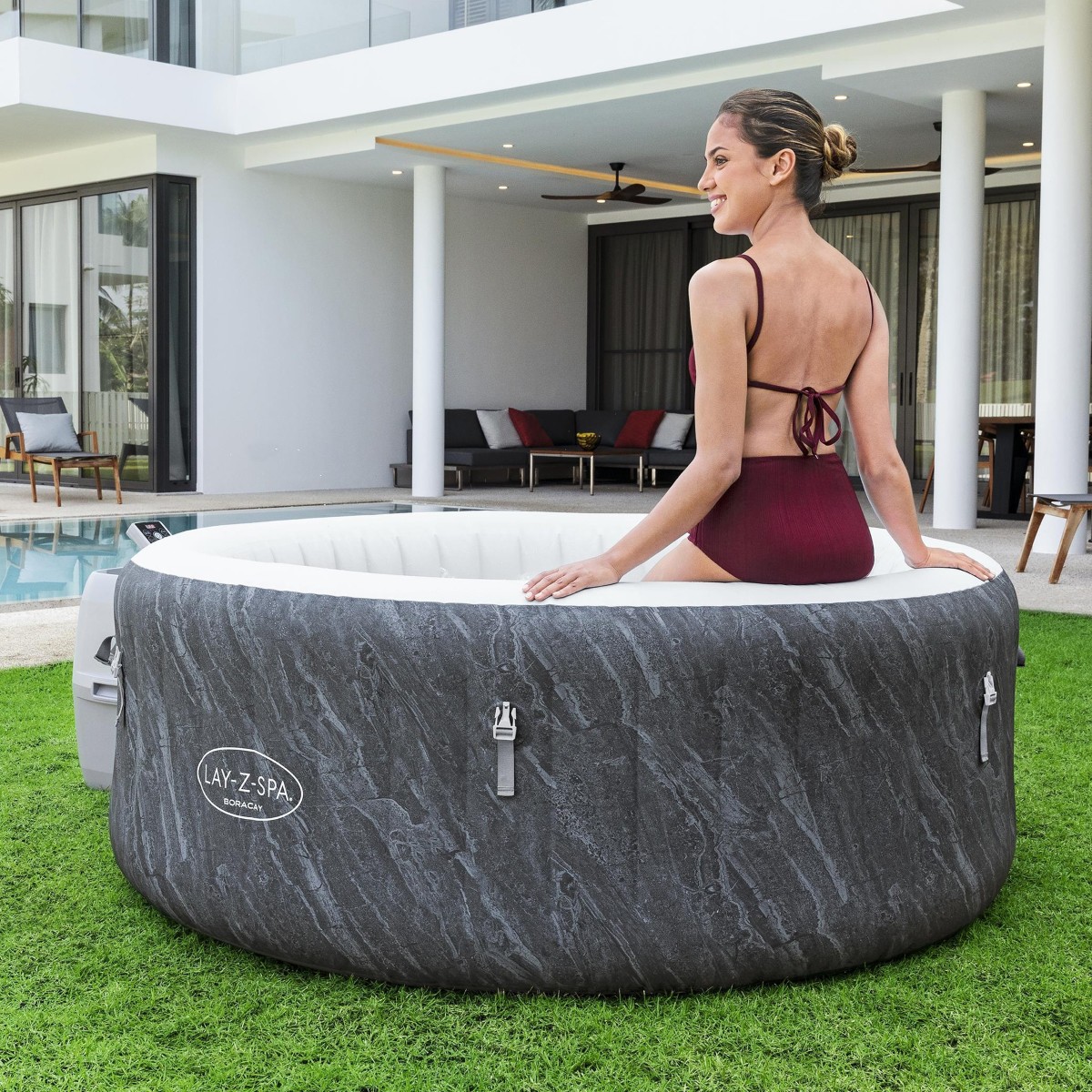 Kobieta w bordowym stroju kąpielowym siedzi na krawędzi jacuzzi Boracay Smart, patrząc w stronę nowoczesnego domu z basenem. Jacuzzi z obudową o wzorze ciemnego marmuru stoi na zadbanym trawniku przed tarasem.